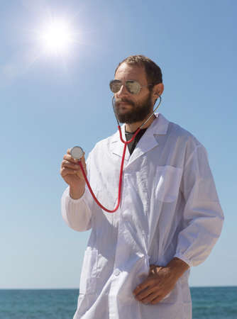 portrait of doctor with stethoscope in ear listen you carefully attentively. American handsome bearded man on blue sky background. Russian Caucasian brutal man in a white coat. Treatment in Israelの写真素材