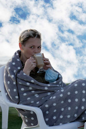 Happy american woman enjoying delicious cup of coffee with milk. Portrait of beautiful Caucasian woman drinking hot coffee wrapped in blanket, against the background of a summer sky with cloudsの写真素材
