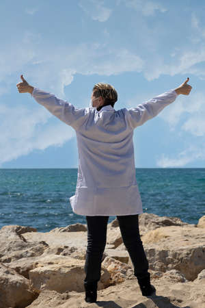 Happy freedom Blond doctor woman with open arms looking at blue sea horizon outdoors with hope. Carefree Russian Caucasian American doctor in medical mask and white coat with back toward cameraの写真素材