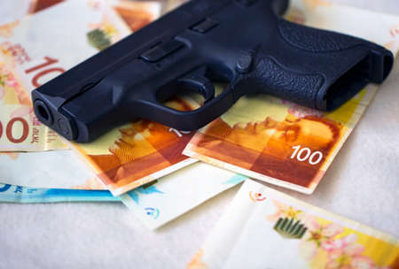 Black gun pistol on stack money Israeli New Shekels banknotes on table. Semi Automatic handgun firearm with New Israel Shekel 100, 200 NIS bills. Currency, criminal money concept. Illegal selling.の写真素材