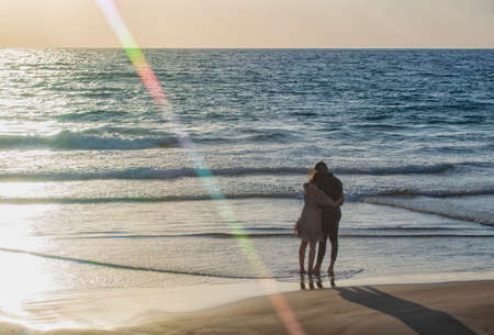 Photo of young happy man kissing and hugging beautiful woman on sunny beach. Loving couple embracing on the sunset with sun's rays and sun reflection in water, enjoying the sunshine. Summer landscapeの写真素材