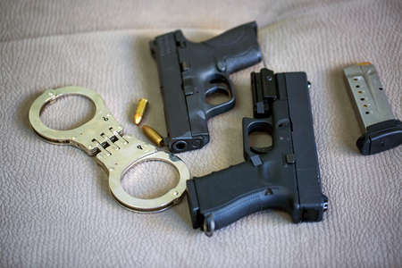 Set of two black guns pistols with ammunition, police handcuffs and bullet shells 9mm. Semi Automatic handgun firearm lie against each other on sofa background. Camera is focused on the handcuffsの写真素材