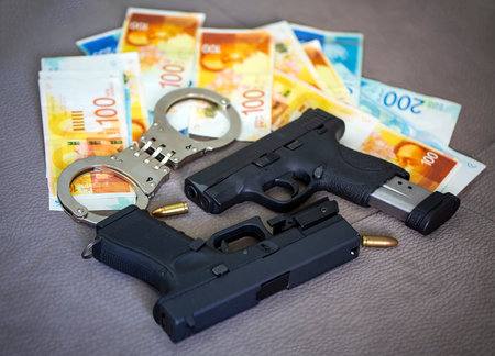 Set of two black guns pistols with ammunition, police handcuffs and bullet shells 9mm with banknotes of Israeli New Shekels. Semi Automatic handguns firearm lie against each other on sofa backgroundの写真素材