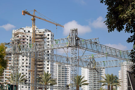 Leaves of a palm tree on a background of high tower crane and residential apartment buildings under construction against blue sky. Real estate development. new haus in framing stageの写真素材
