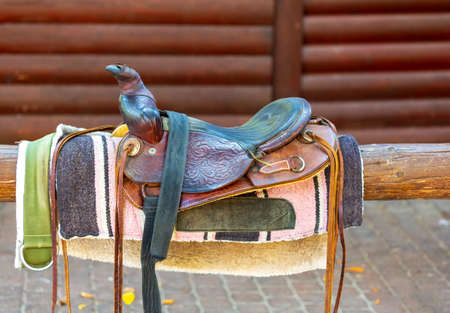 Old saddle on fence against the wooden wall. Sport leather saddle, harness for horses on daylight. Western horse saddles on a rack, ready for dressage training. Equestrian sport background, vintage.の写真素材