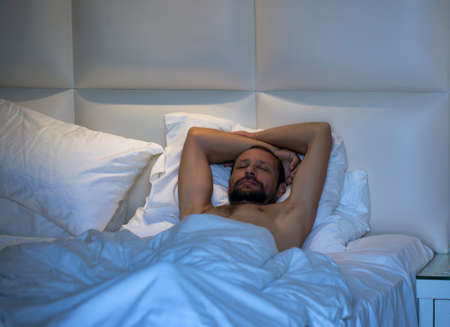 Sleepy bearded man lying in bed on his back under a white blanket with his hands behind his head....