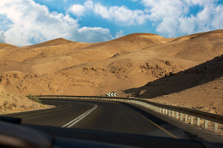 Front view through car's window on bendy road and mountain fantasy landscape on blue sky background with clouds. Sunny sky over cliffs, large salt mountains Sodom and Gomorrah. Judean desert, Israelの写真素材
