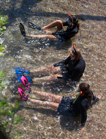 Three female divers with scuba masks on sitting in shallow waters wearing their swim fins, preparing to dive into the Red sea. Divers sits on the shore and puts on a scuba weight, flippers and a maskの写真素材
