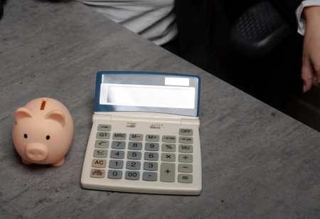 This image shows cute pink piggy bank with calculator. Saving money wealth and financial concept. Close-up view of money box on desk in office. Coin bank with calculating machine on wooden table.の写真素材
