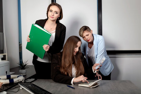 Three female colleagues at a work laughing while working at an office table. Female with long hair holding bundle dollars and counting revenue. Womans with cash money in dollar banknotes in hand.の写真素材