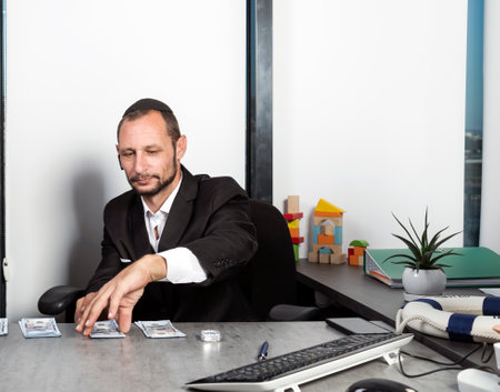 Serious jewish man in formal wear sit at desk wearing kippah with surprised looking at dollar bills. Caucasian man is putting hundred dollar bills on the table as payment. Jew receiving salary moneyの写真素材