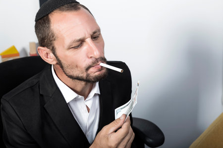 Handsome Bearded Jewish man in Yarmulke (hat, Kippah) and formalwear lighting a cigarette using hundred dollar banknote. Brutal jew man or businessman with beard lighting cigare from burning banknoteの写真素材