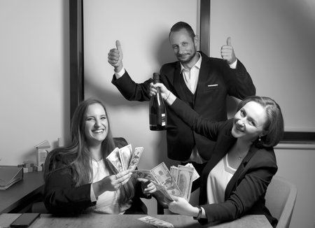 Black and white photo of two happy women sitting at desk looking at camera holding a bundle of money and champagne bottle celebrating the big event. Happy excited bearded man smiling with a fingers upの写真素材