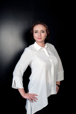 Businesswoman with hairstyle in elegant white shirt standing with hands on thighs and posing against black wall. Young beautiful cheerful brunette caucasian female with dark brown hair. Lovely girlの写真素材