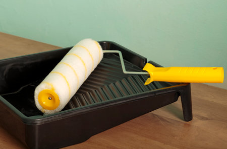 Paint roller with a yellow handle and a black plastic paint tray on wooden board. Building, renovation and construction work. Tools for painting walls: roller, tray near a green wall. interior updateの写真素材