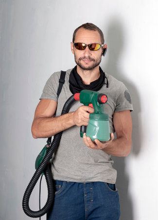 Caucasian bearded men with sunglasses holds green spray gun in his hands. Repair of apartments, houses and paints the wall concept. Workers hold paint gun holding by mans hand on white wall backgroundの写真素材