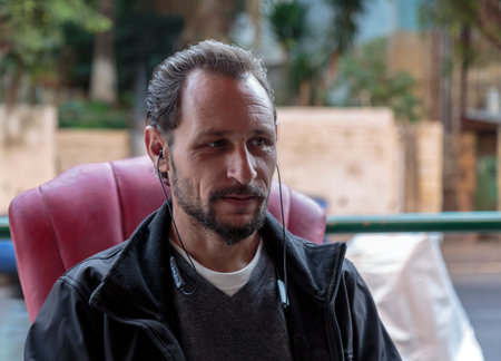 Caucasian man sitting looking away to side with natural expression. Handsome old jewish bearded man with a receding hairline with earphones sitting on a red chair wearing a simple black jacketの写真素材