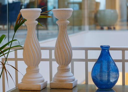 Single blue vase, two white candle holder stand on the table as part of table decoration, decor. Large decorative candlesticks on a stand against the background of the white wall and green plantの写真素材