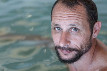Happy adult caucasian man with beard and mustache looking at camera. Close up portrait of man's face With Water Drops On Smooth Skin against blurred water background in the pool with space to copy.の写真素材