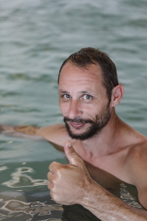 Happy adult caucasian man show thumbs up with beard, mustache looking at camera. Man's face With Water Drops On Smooth Skin against blurred water background man shows ok in the pool, selective focusの写真素材