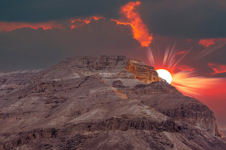 Sunset against backdrop of mountains and fiery red sun in the dark sky in Dead Sea, Israel. Beautiful desert landscape among the sands and stones. Large stone ridge with dramatic clouds and sun raysの写真素材