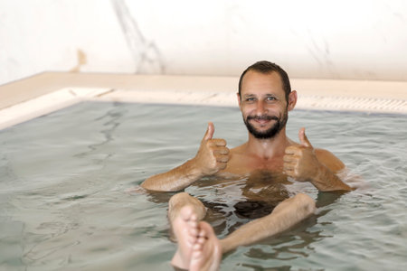 Adult caucasian man with beard, mustache floating in the swimming pool show double thumb up looking at camera. Bearded man with water drops on skin against blurred water background shows two ok, yesの写真素材
