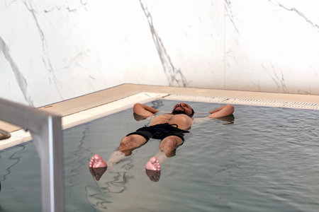Adult caucasian man with beard, mustache floating in the swimming pool on his back with clear blue water. Bearded man with his hands under his head enjoying in the pool. Guy lying in water on his backの写真素材
