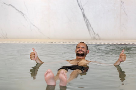 Adult caucasian man with beard, mustache floating in the swimming pool with dense salt water and show double thumb up, ok, yes. Bearded man floating in the spa pool with water from the Dead Seaの写真素材
