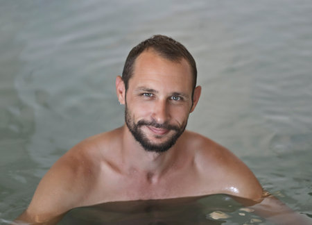Happy smiling adult man enjoying indoor swimming pool. Caucasian man with beard, mustache floating in the swimming pool with dense salt water. Portrait of handsome bearded guy relaxing in the spa poolの写真素材