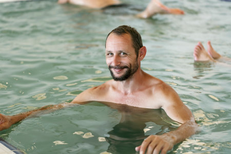Happy mid adult, middle aged caucasian guy smiling in the indoor swimming pool with dense salt water, Dead Sea. Bearded man relaxing, enjoying in the spa pool. Positive delighted man sitting in waterの写真素材