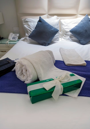Luxurious bedroom with two towels on the bed. Double bed in hotel room. Big comfortable white bed in sleeping room. Interior with bed lines. Blue pillows and green gift on bed with white headboardの写真素材