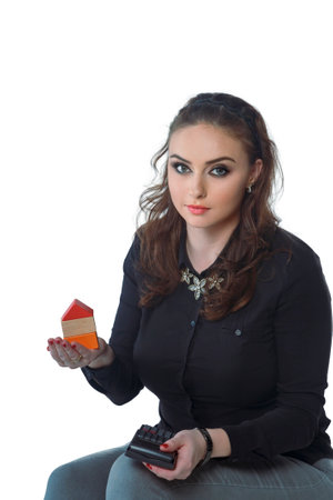 Young beautiful woman, businesswoman worker or manager sitting at her work place isolated on white background holding construction blocks, miniature house, toy house in hand and using a calculator.の写真素材