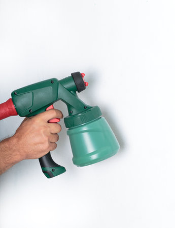 Hand holding a green paint sprayer against white wall background. A close up of a hand gripping a green electric paint sprayer, ready for use. A person, man using a spray gun for home renovation.の写真素材