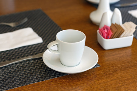 A white coffee cup rests on a saucer, accompanied by cutlery wrapped in a napkin. A dark woven placemat lies underneath the cup, a white holder containing various colored sugar packets. dining tableの写真素材