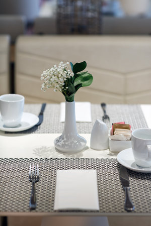 The focus is on the center of the table where a white vase with blooming white flowers is placed, surrounded by dining ware including cups, saucers, and utensils. A small container holding of sugarの写真素材