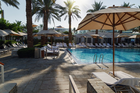 Bright sunlight, creating a warm and inviting atmosphere. White lounge chairs are neatly arranged around the pool. Some of the chairs are shaded by beige umbrellas, offering relief from the sun.の写真素材