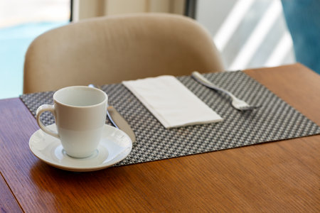 A white cup and saucer sit on a woven placemat on a wooden table. Beside them, a folded white napkin and silver fork are neatly placed. The chair in the background and natural light through the windowの写真素材
