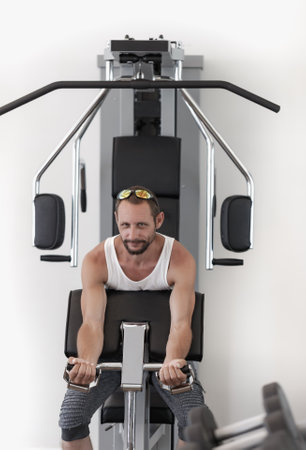 An individual is using an indoor rowing machine. Dressed in a white top and dark shorts, the person is mid-motion, pulling the handle of the black and silver machine, positioned against a white wallの写真素材