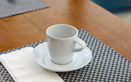 An empty white cup on a saucer, resting on a woven placemat. A folded white napkin is beside the saucer. The table surface is wooden. The coffee mug, to come, sit, and enjoy their own moment of rest.の写真素材