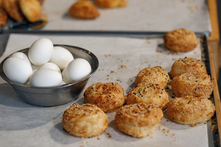 A baking sheet with golden brown rolls sprinkled with seeds. Next to the rolls, there's a bowl of white eggs. Israeli breakfast including baked pastry bourekas and egg. A focus on the rolls and eggsの写真素材