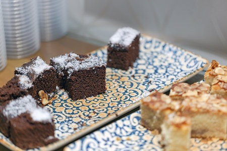 Tray of desserts, brownies topped with powdered sugar and almond slices. A cake with almonds is also visible. The tray has a blue and yellow pattern. In the background, there are stacked of cupsの写真素材