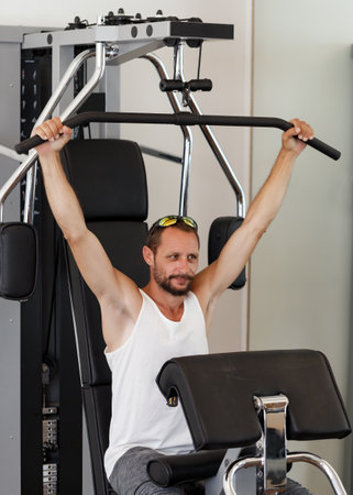 A man using a gym machine pulling down a bar attached to weights, wearing a white tank top and gray pants showing muscular arms. The machine is black and silver located indoors gym with light wallsの写真素材