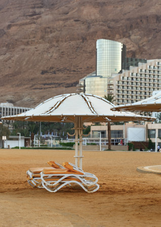 White umbrellas and lounge chairs are prominent in the foreground, placed on sandy ground for relaxation. The background contrasts with a large rugged mountain under a clear sky. Dead Sea, Israelの写真素材
