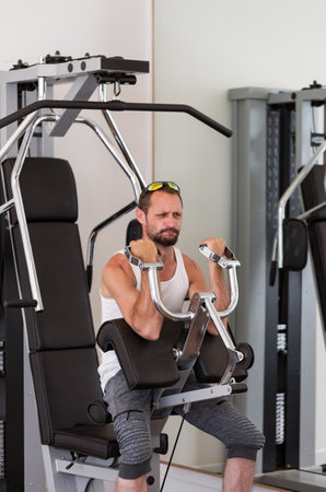 A man exercising in a gym. Male using a machine for upper body workouts and they are wearing sunglasses a white tank top, and gray shorts. The gym appears well-equipped. Strength and motivation, sportの写真素材
