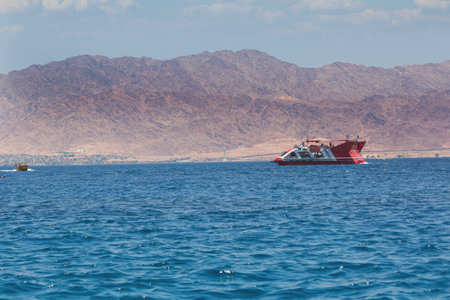 A red and white boat floats on calm blue waters. In the background, rugged brown mountains contrast beautifully with the serene sea. The sky is clear, adding to the overall peace of the scene.の写真素材