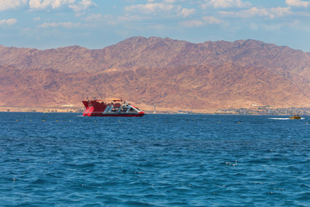 Red and white modern boat sailing on a tranquil blue sea. The backdrop features rugged mountains under a clear sky. The mountains, though barren, add a majestic element to the scene. Red Sea, Israelの写真素材