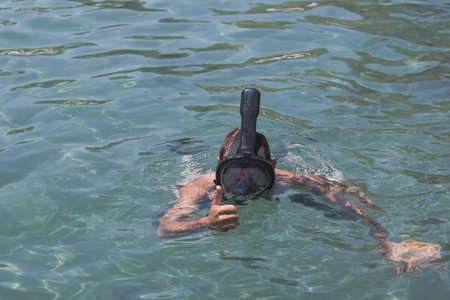 A man wearing a full face snorkel mask gives a thumbs up while swimming in the sea. Clear blue waters. With a gesture of approval, he shows his enjoyment of the summer vacation and snorkeling activityの写真素材