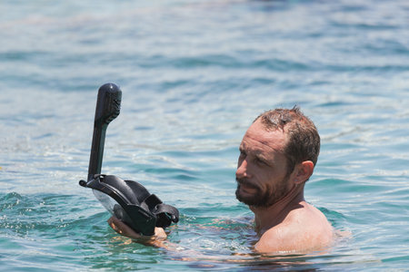 Man holding black snorkel mask while swimming in clear blue ocean during daytime. A man with a beard is partially submerged in sea water, holding a black snorkel mask. Sunlight reflects off the wavesの写真素材