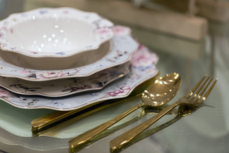 Stack of porcelain dishes with golden cutlery on a glass table. Below the bowl are plates of increasing sizes with floral designs. A golden spoon and fork are placed beside the stacked dishes.の写真素材