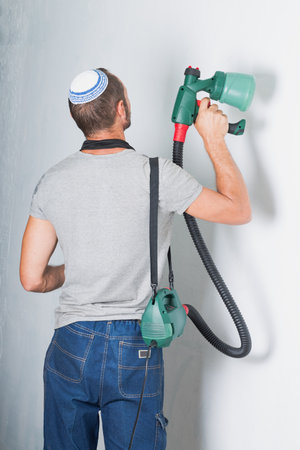 Jewish man using a paint sprayer for home renovation. He is seen from behind, wearing a kippah, gray shirt, and blue jeans. In his hands holds green sprayer with a hose attached, connects to a deviceの写真素材